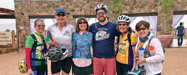 Two men and four women dressed in bicycling clothes and helmets stand in front of a brick wall in a sandy location